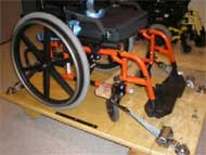 This figure shows three pictures of  a) manual wheelchair secured by 4-point strap-type tiedown; b) top view of UDIG auto docking system;  c) powered wheelchair in rear-facing passenger station (RF-WPS) 